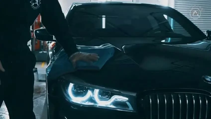 A worker's hand wiping the bonnet of a dark car with a microfibre cloth, headlights on